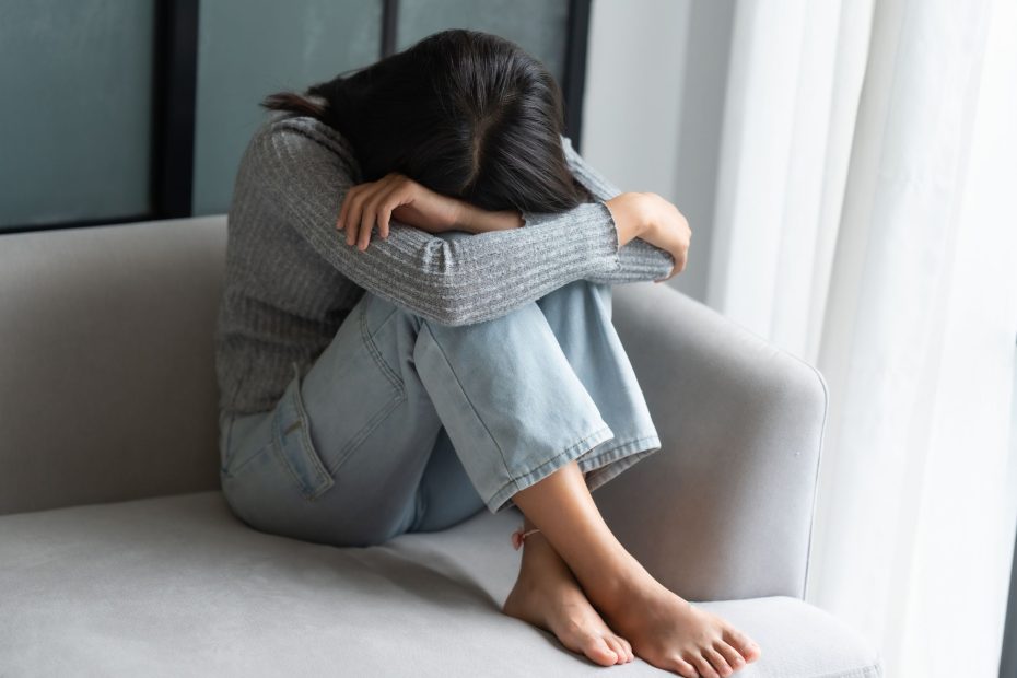 Woman sitting on couch with knees bent up, arms resting on them, and face hidden.