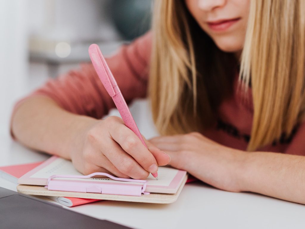 Girl writing with a pink pen in a pink notebook.